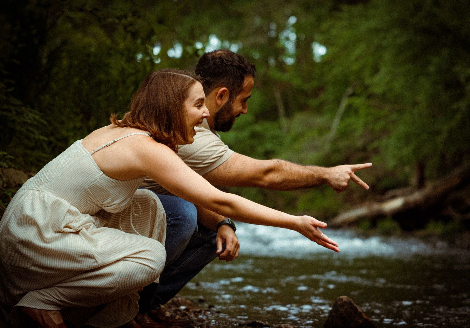 Two people in casual attire crouch by a forest stream while pointing at something in the water.