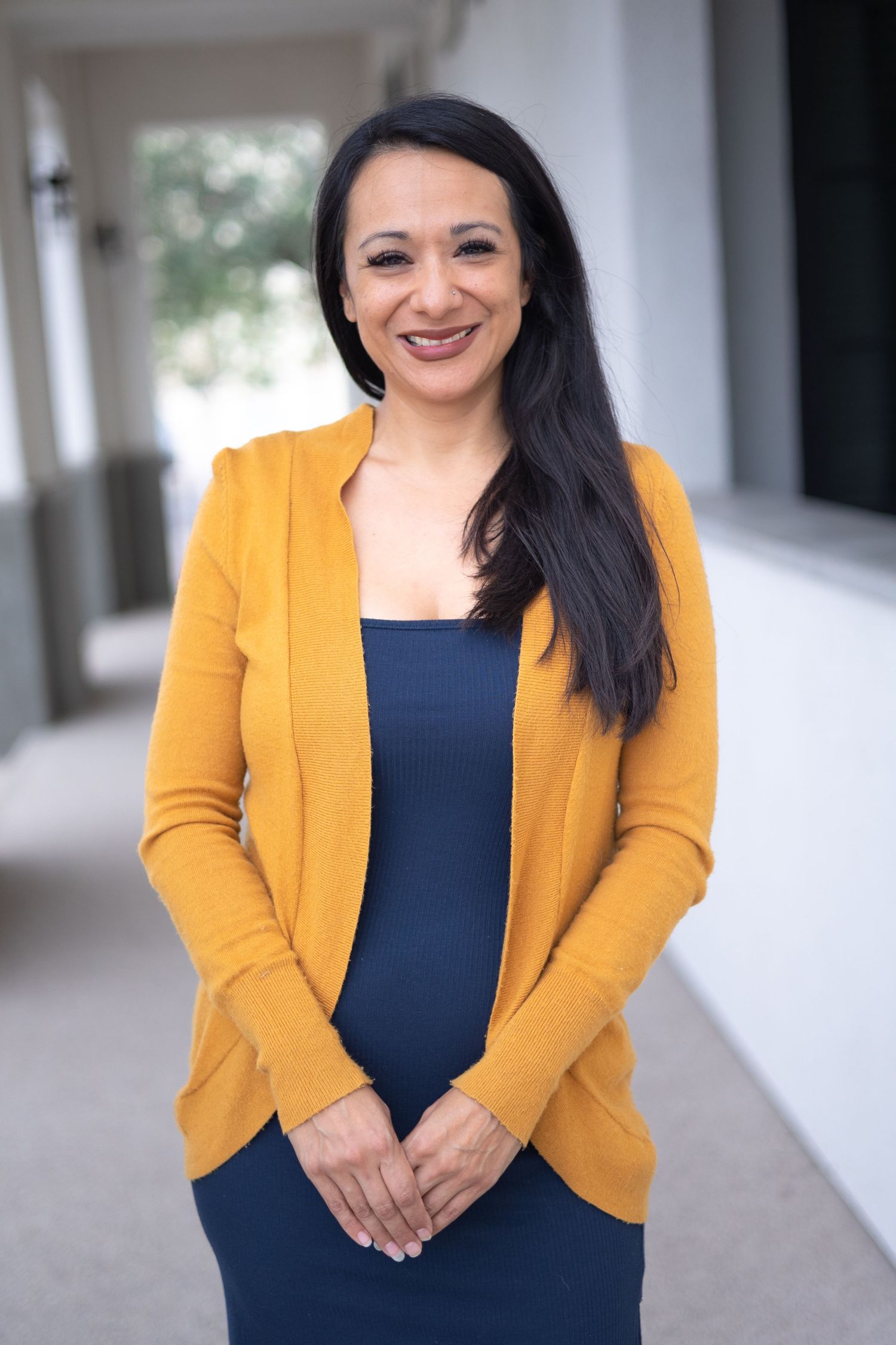 Professional portrait of a woman wearing a mustard yellow cardigan over a navy blue dress, standing in a bright corridor in Dallas