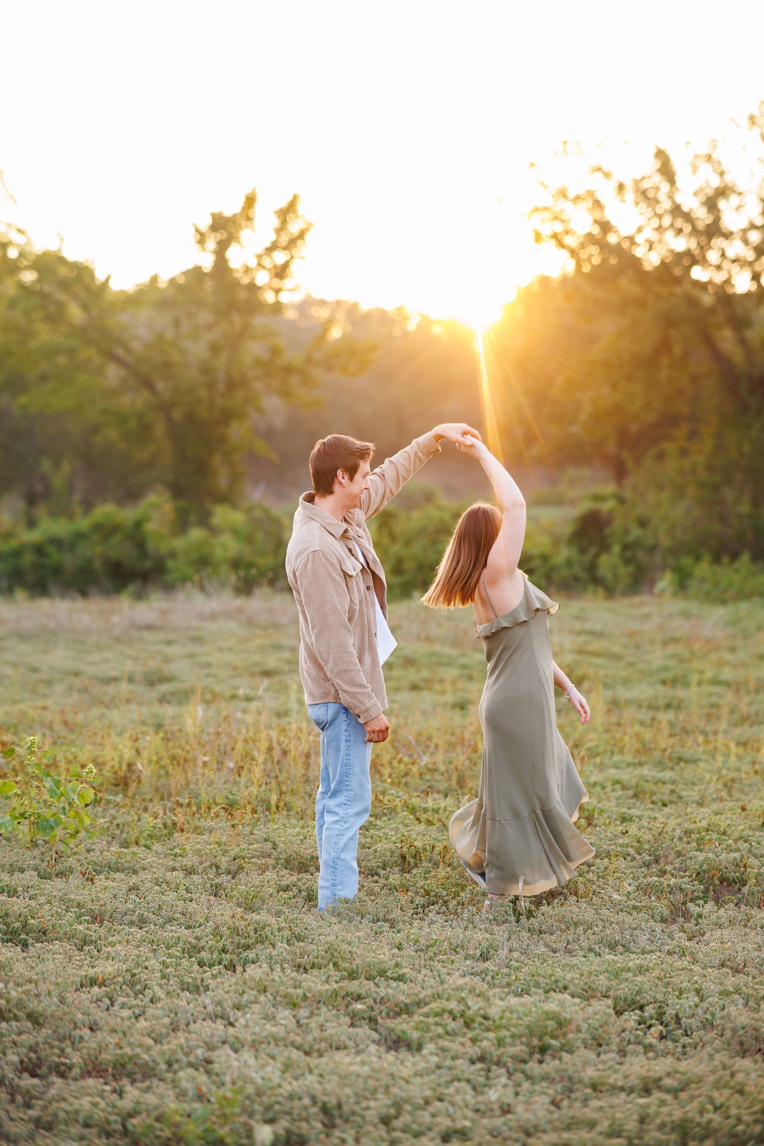 Couple dancing together in a sunlit meadow during golden hour as the woman twirls in her long dress, during their Dallas engagement session