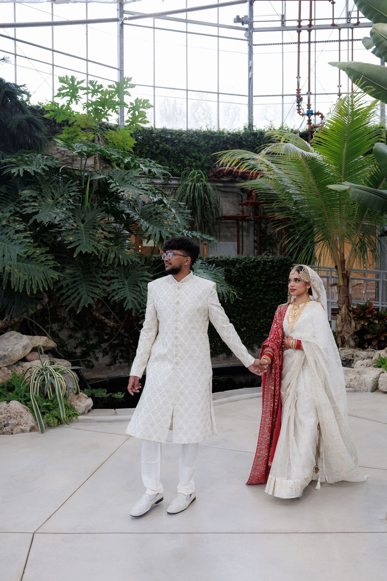 Newlyweds in traditional Indian wedding outfits posing in botanical garden setting.