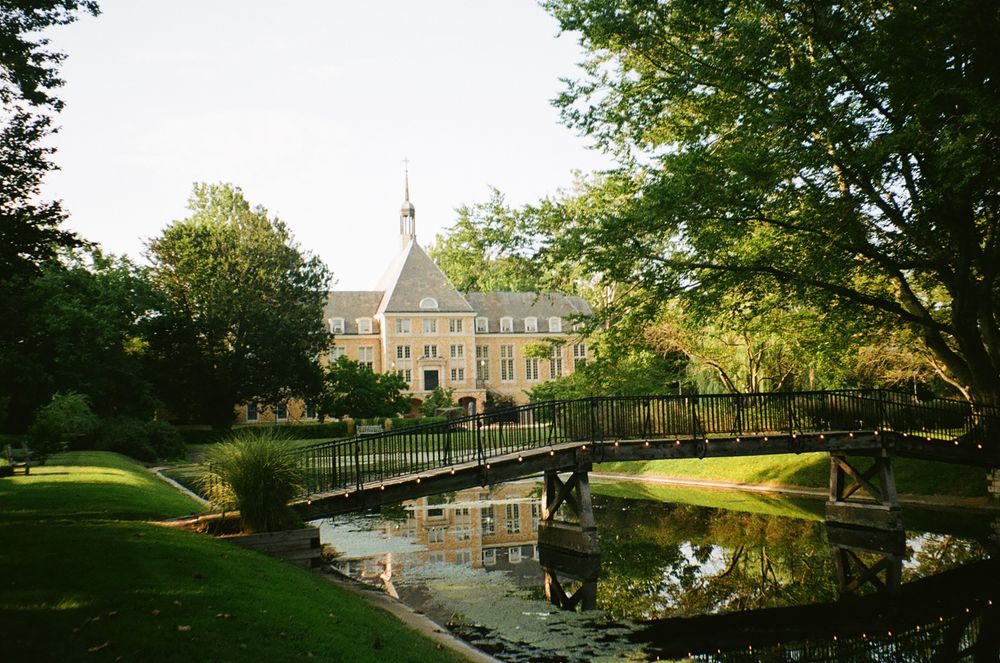 Elegant manor house with a pond and wooden bridge in a lush garden setting during golden hour.