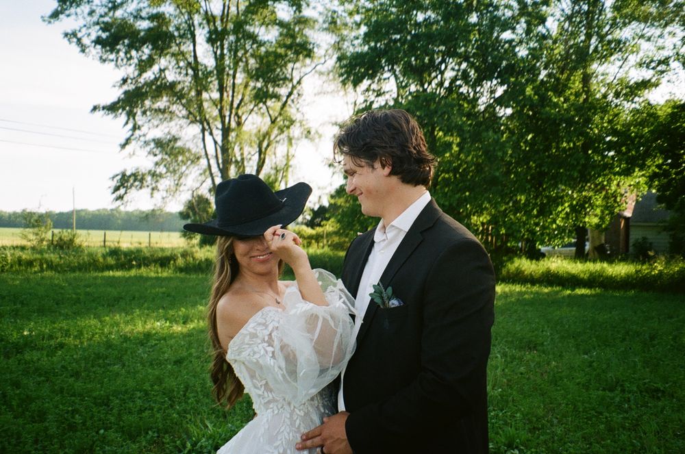A couple shares an intimate moment outdoors in a sunlit field with trees, the bride wearing a white dress and black hat.