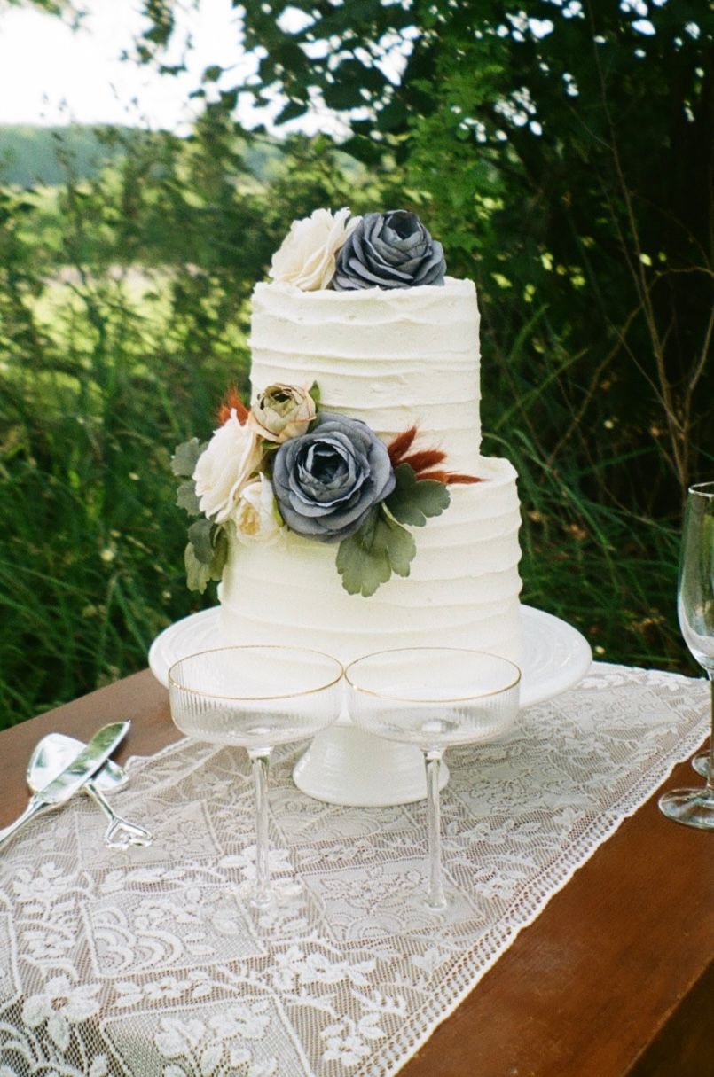 Elegant three-tier white wedding cake decorated with gray and white roses sits on lace tablecloth outdoors.