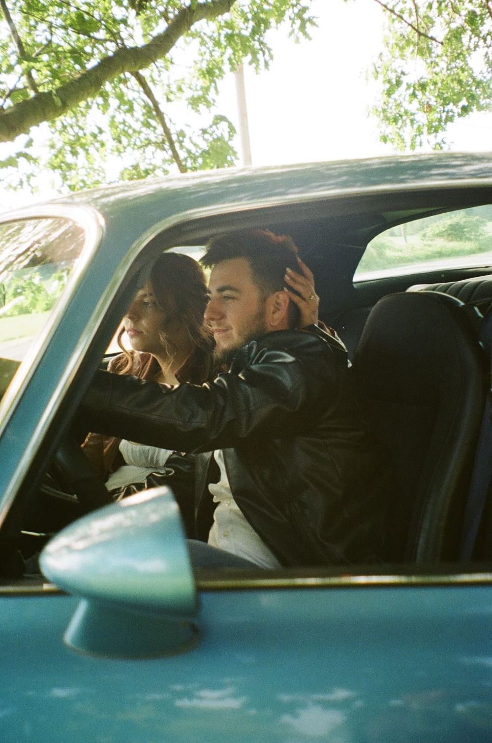 Two people seated in a turquoise car with sunlight filtering through trees in a candid outdoor shot.