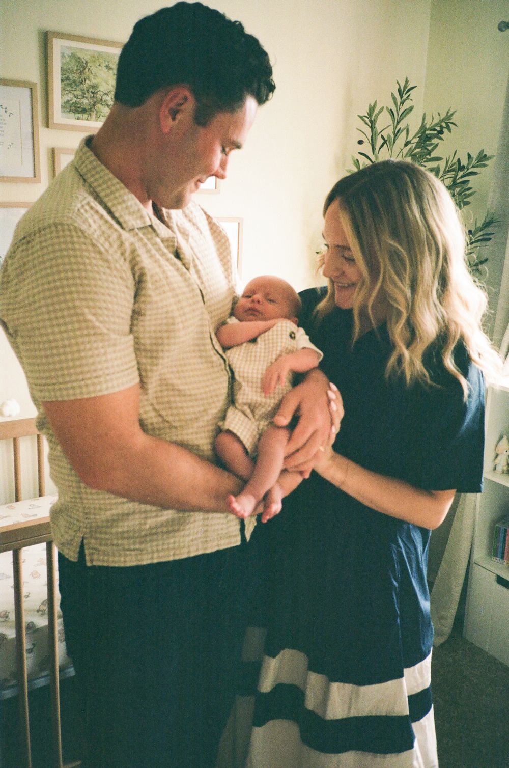 A couple looks lovingly at their newborn baby together in a sun-lit room with indoor plants.