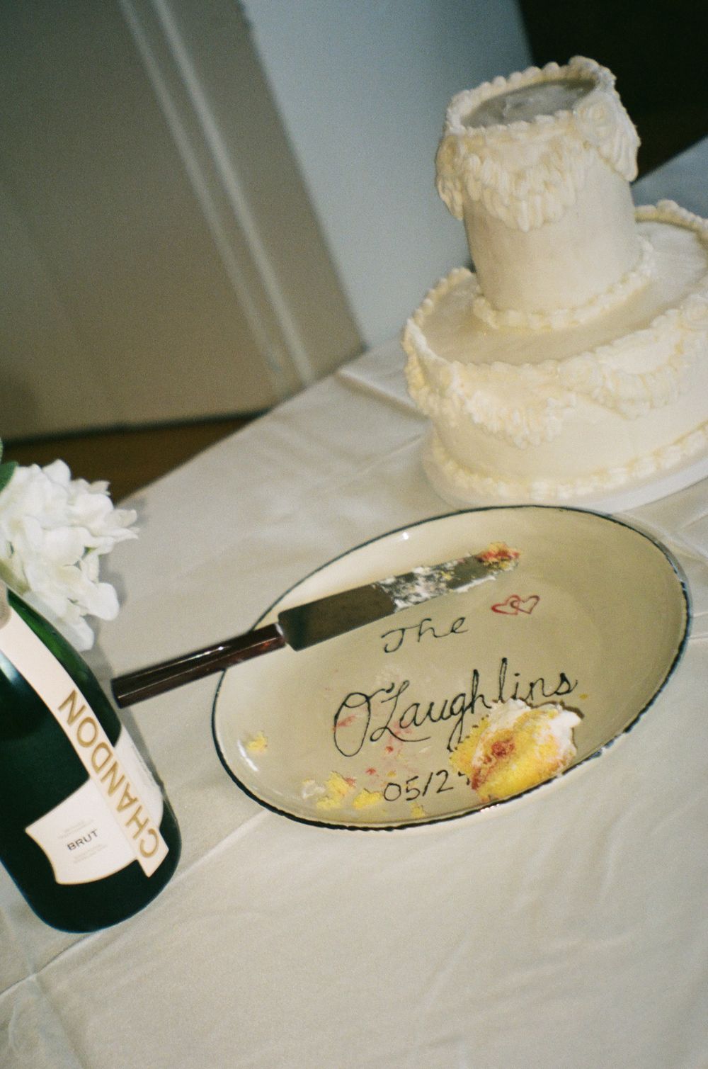 A white wedding cake and champagne bottle sit beside a decorative serving plate with cursive writing and floral design.
