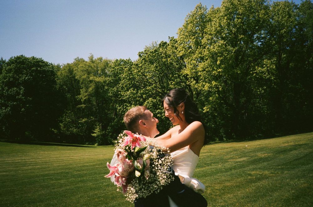 A newlywed couple share a romantic moment in a sunny meadow with a bouquet of flowers.
