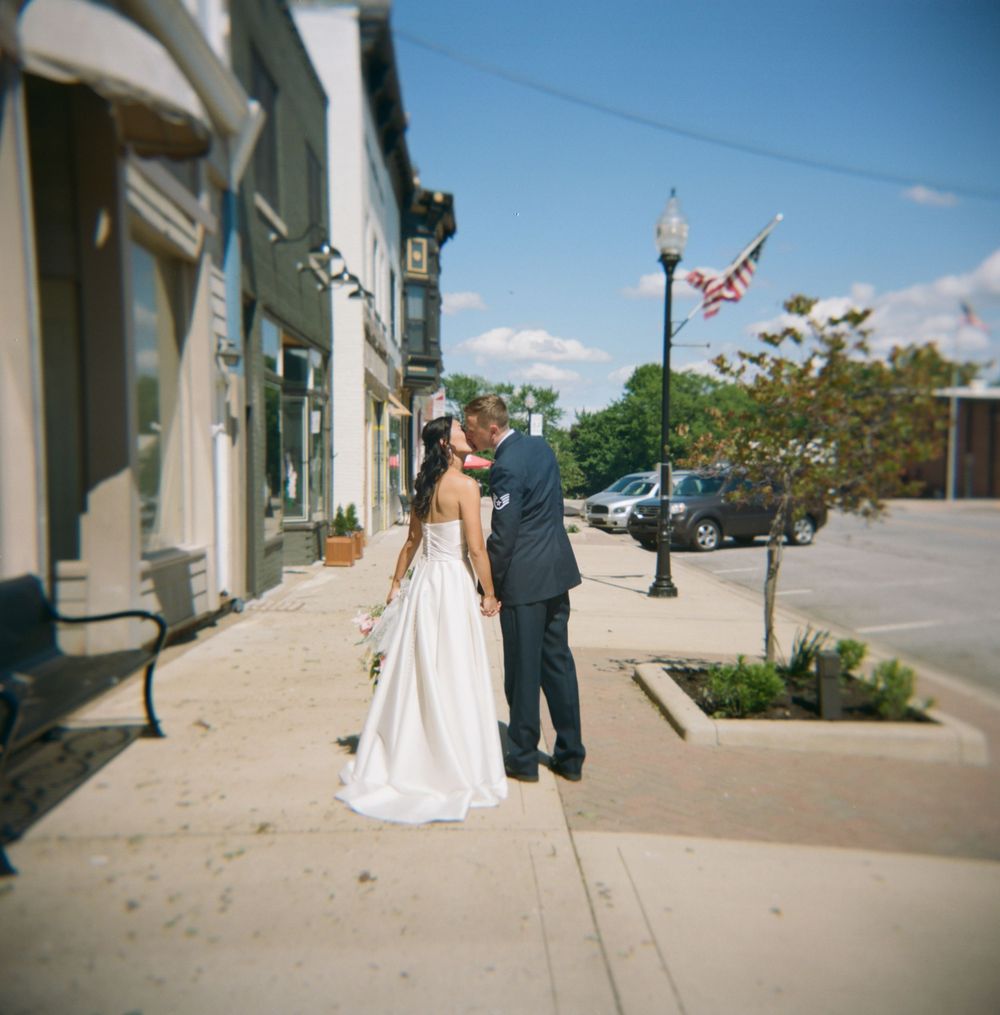A bride and groom share a romantic moment on a sunny sidewalk in front of storefronts and vintage lampposts.