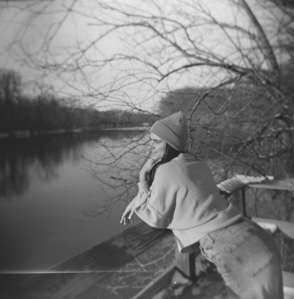 A figure in winter clothing leans against a railing overlooking a serene lake with bare tree branches in the foreground.