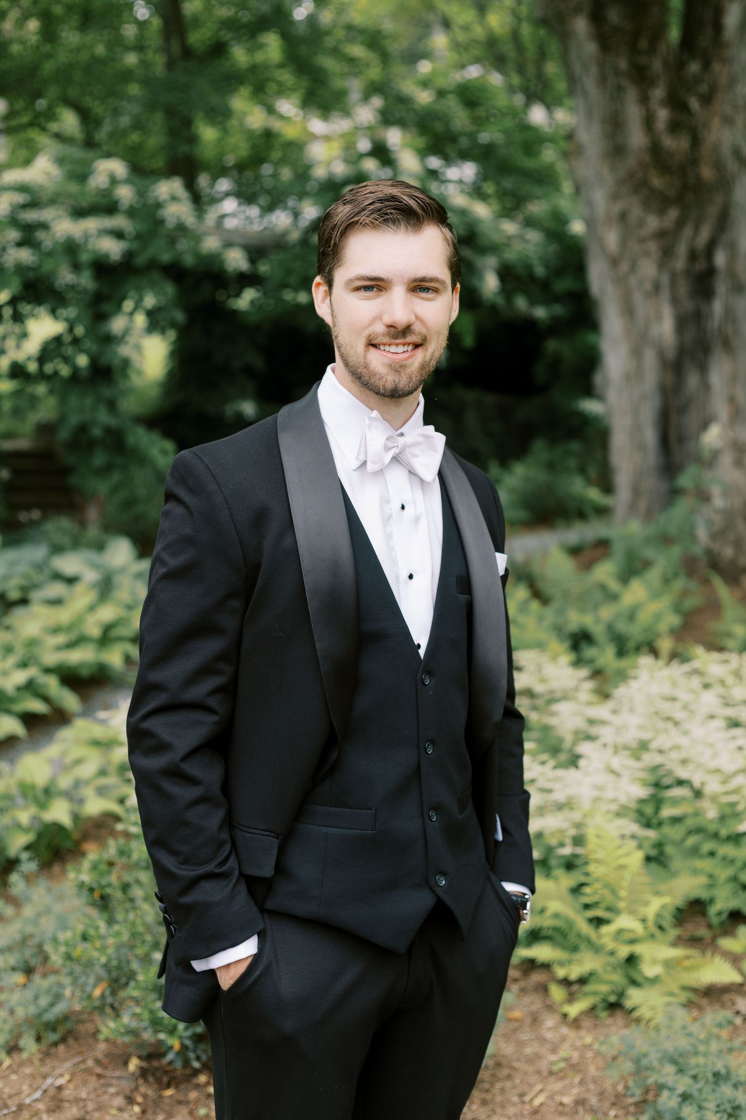 Well-dressed person in formal black tuxedo and white bow tie poses confidently outdoors among lush greenery.