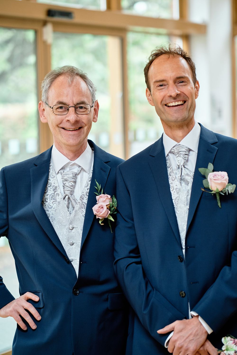 Two men in matching navy suits and silvery paisley ties wearing pink rose boutonnieres smile at a formal event.