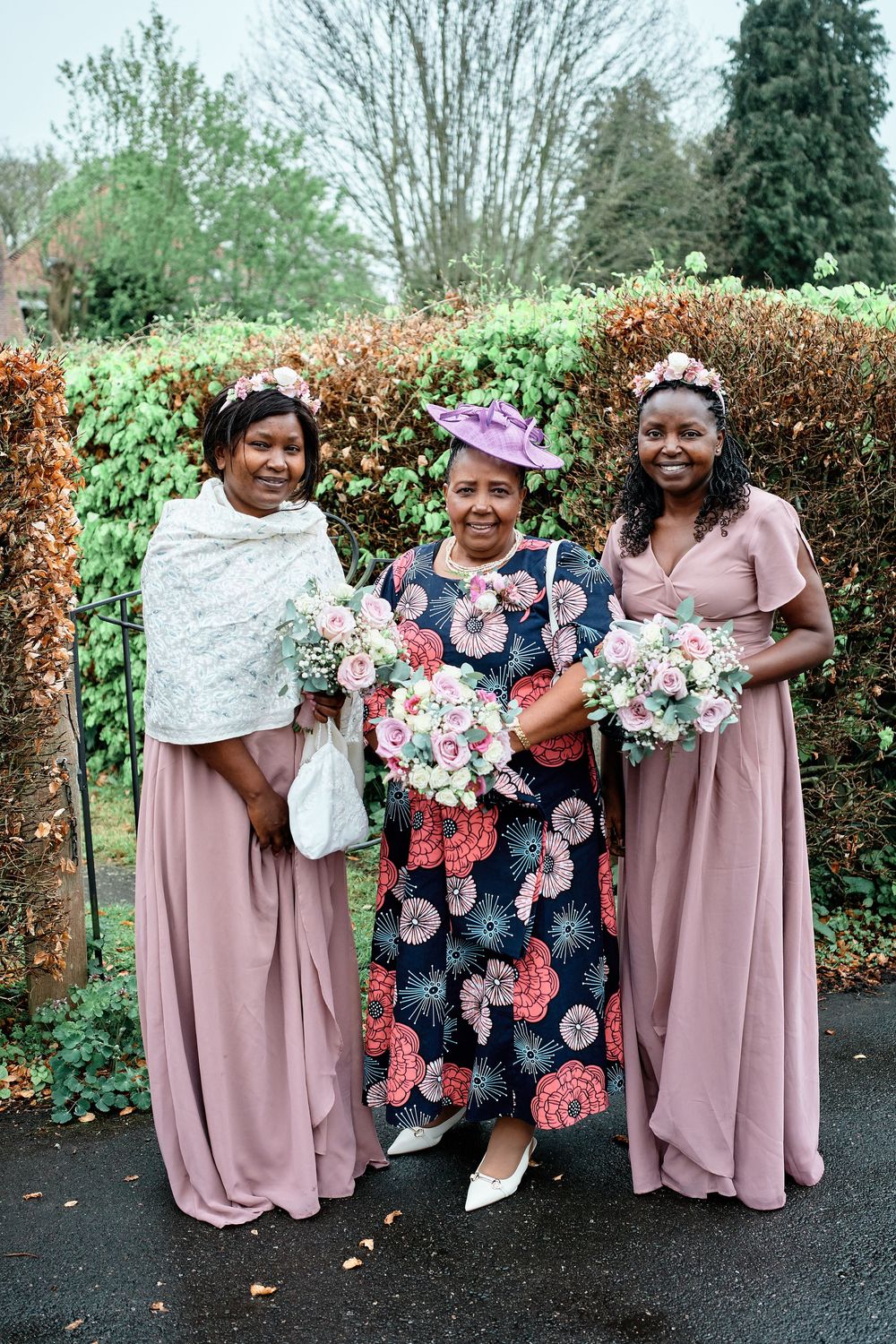 Three people wearing formal attire stand together in a garden setting holding pink and white floral bouquets.