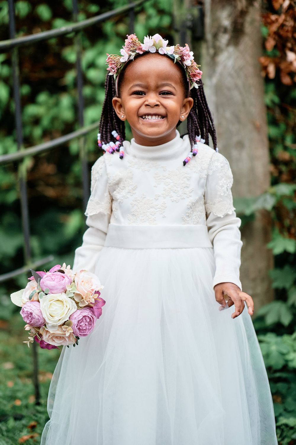 Young attendant in white dress holding pink rose bouquet with flower crown in outdoor garden setting.