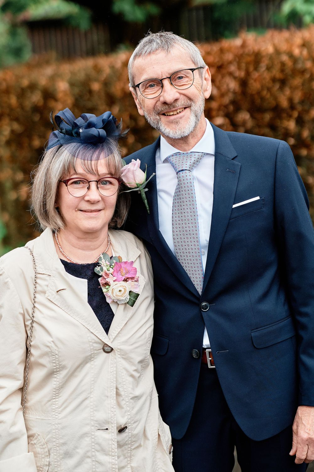 A couple poses outdoors at a formal event, wearing a navy suit and beige jacket with floral corsages.