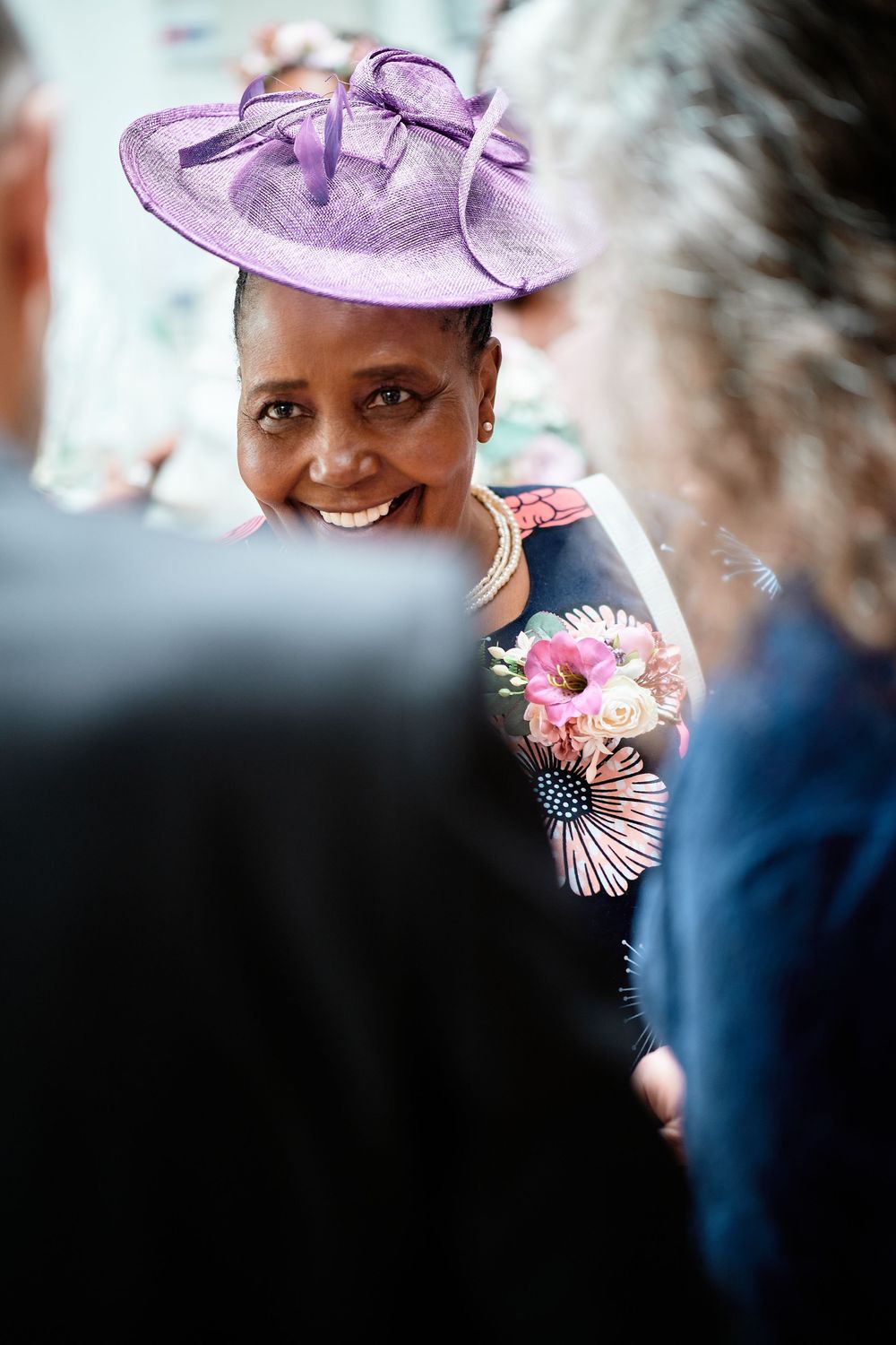 A figure wearing a lilac purple hat and floral dress smiles warmly while attending a formal event.