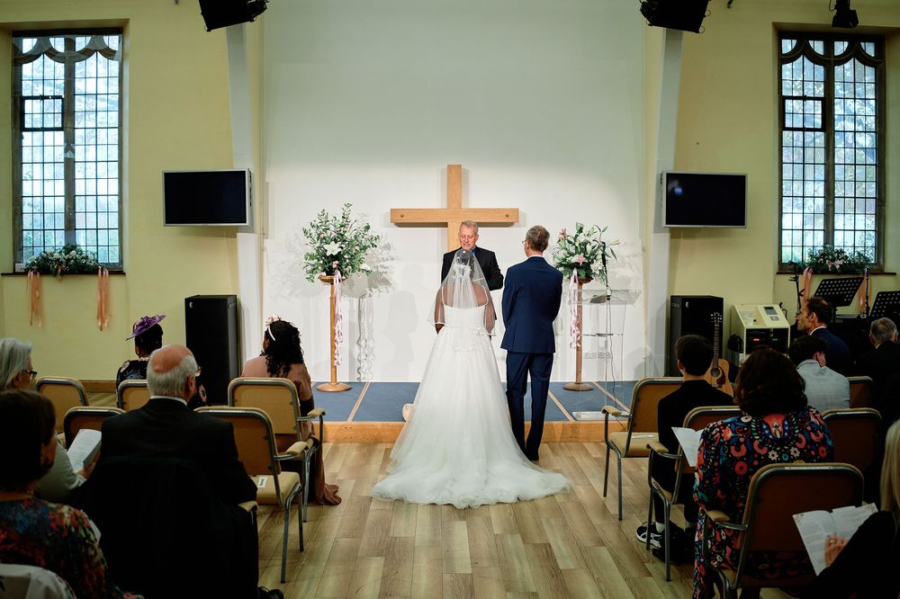 Wedding ceremony in a traditional chapel with wooden floors, stained glass windows, and floral arrangements by the altar.