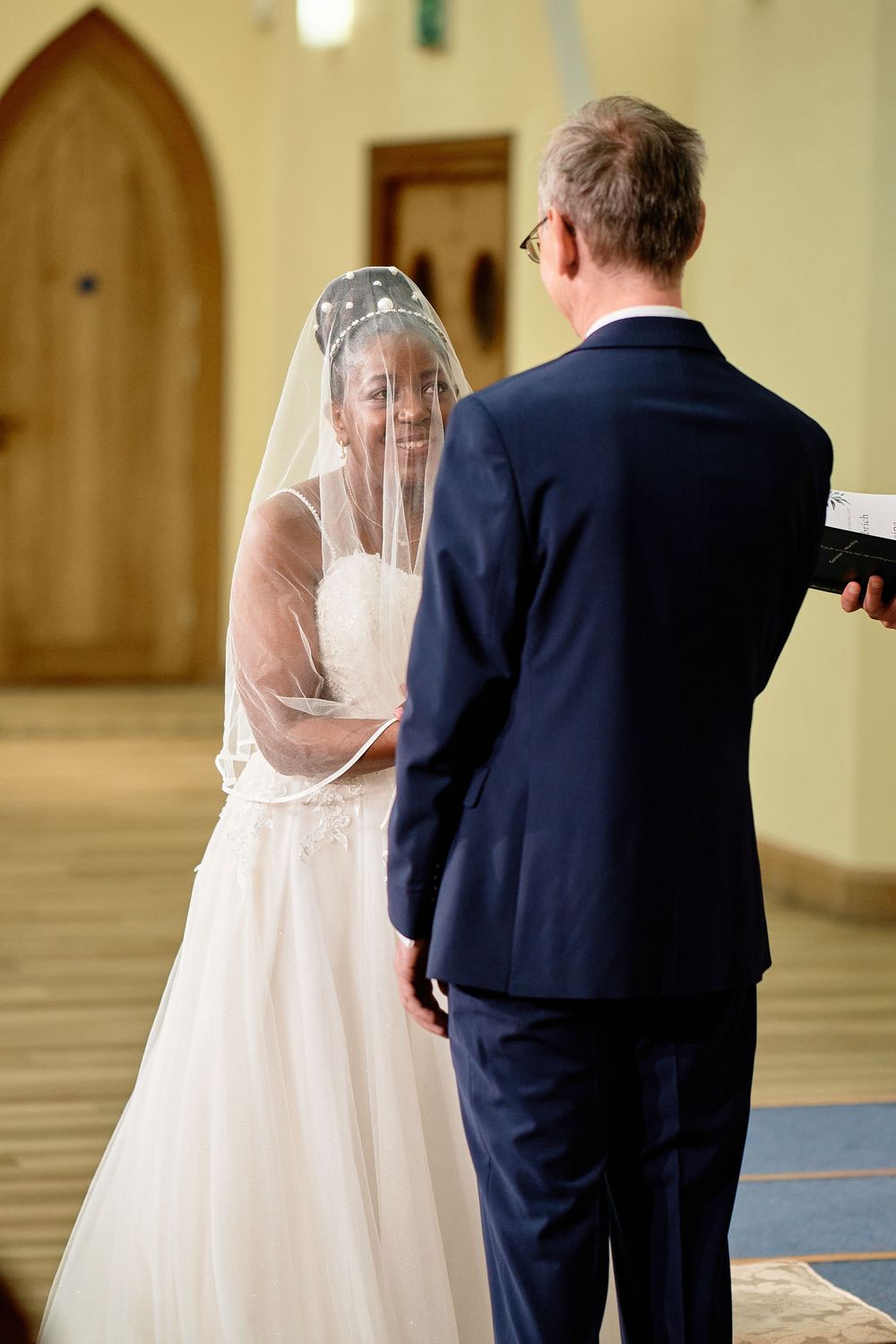 Bride in white wedding dress with veil stands across from groom in navy suit during indoor church ceremony.