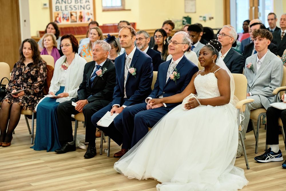 Wedding guests sitting in rows of chairs at an indoor ceremony with wooden floors and sunlit windows.