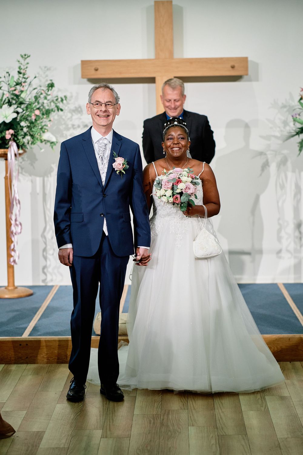A couple stands together at a wedding ceremony in front of a wooden cross, with the bride in a white ball gown.
