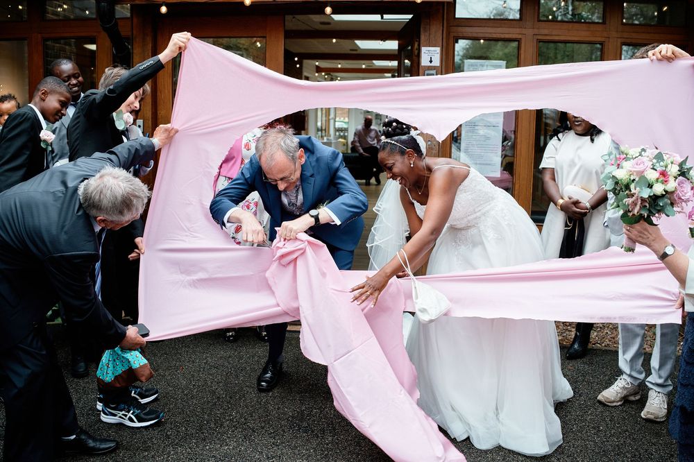Newlyweds duck under a pink fabric held by wedding guests during a traditional wedding game or celebration.