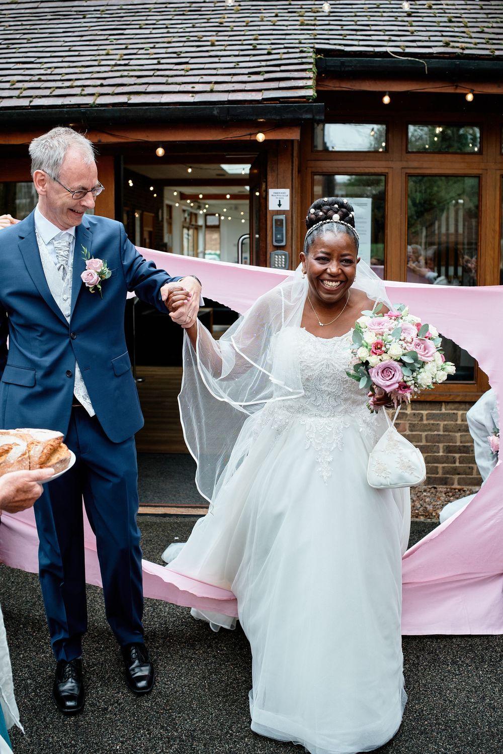 A joyful newlywed couple celebrates outside a venue, with the bride in a white dress holding a pink and white bouquet.