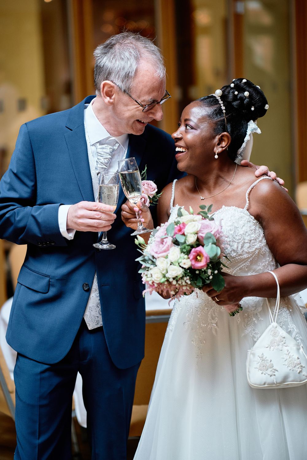 A bride in a white wedding dress and groom in a navy suit share a joyful moment while toasting with champagne glasses.