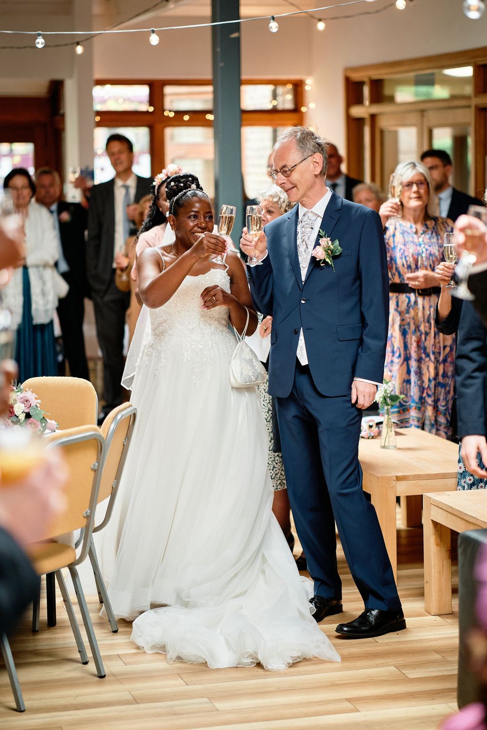 Two newlyweds share a joyful toast at their wedding reception while guests look on in an elegant indoor venue.