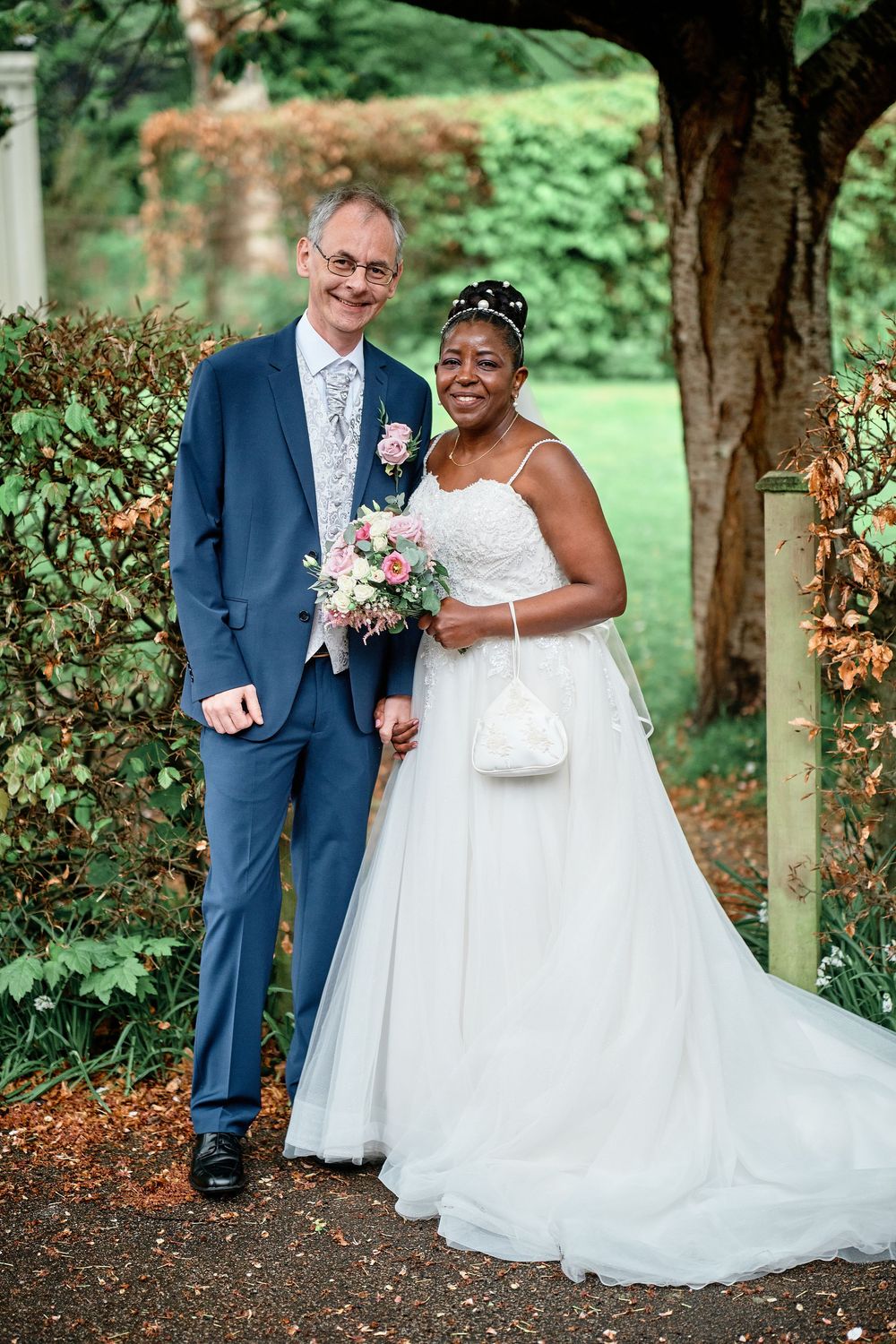 Newlywed couple poses together in a garden setting with bride in white dress holding pink bouquet and groom in navy suit.