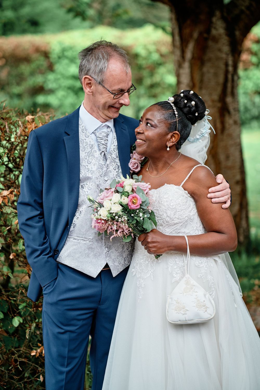 A bride and groom share a joyful moment outdoors, with the bride holding a pink bouquet and wearing an elegant white gown.