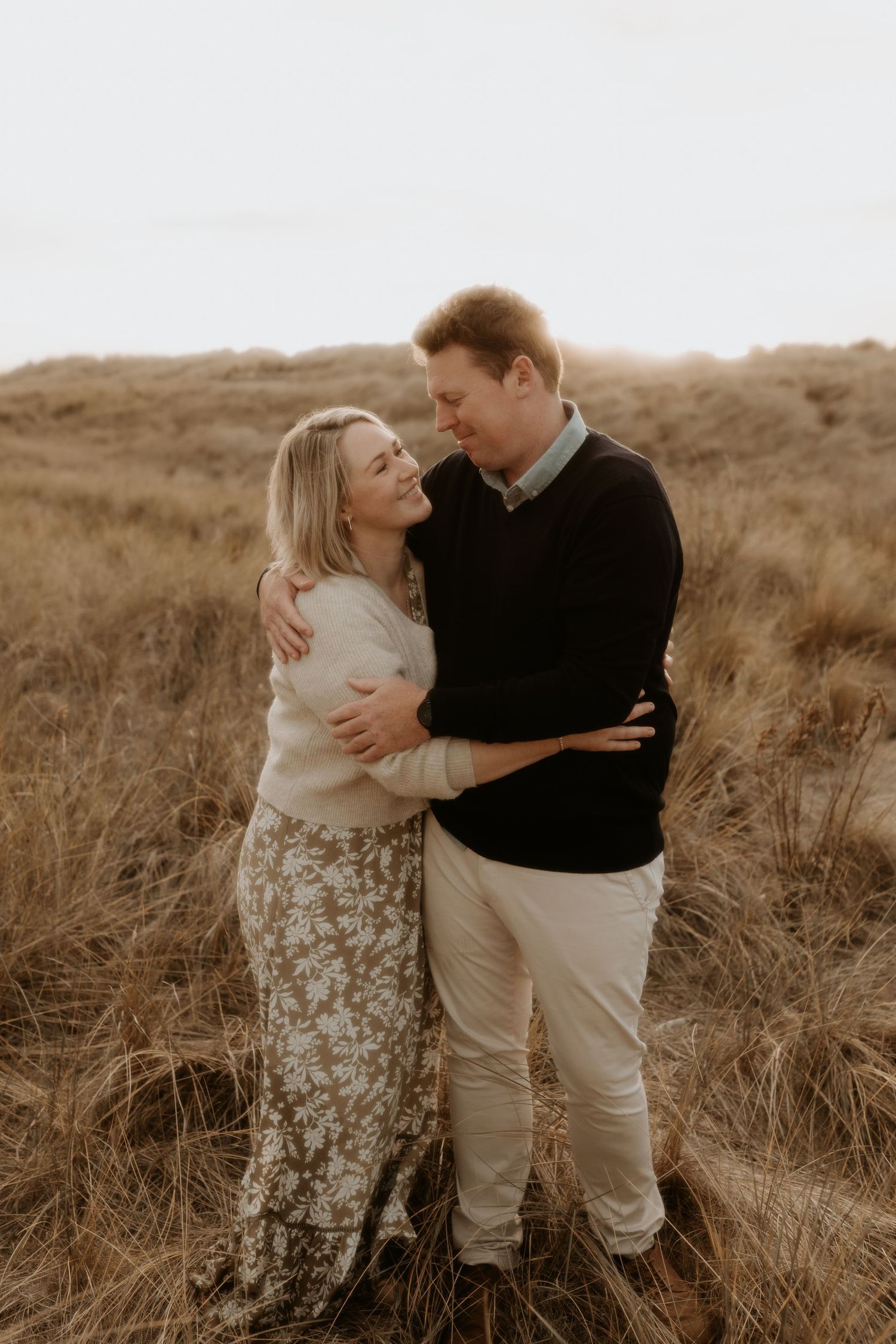 A couple embraces in a romantic pose among tall grass during golden hour in a rustic outdoor setting.