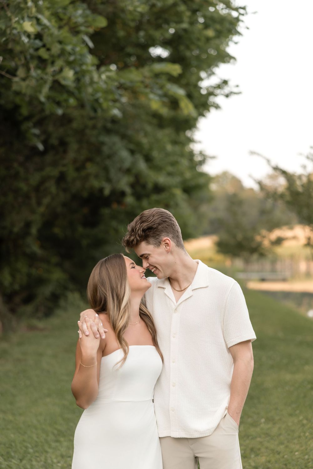 A romantic couple shares an intimate moment together on a grassy lawn near trees at sunset.