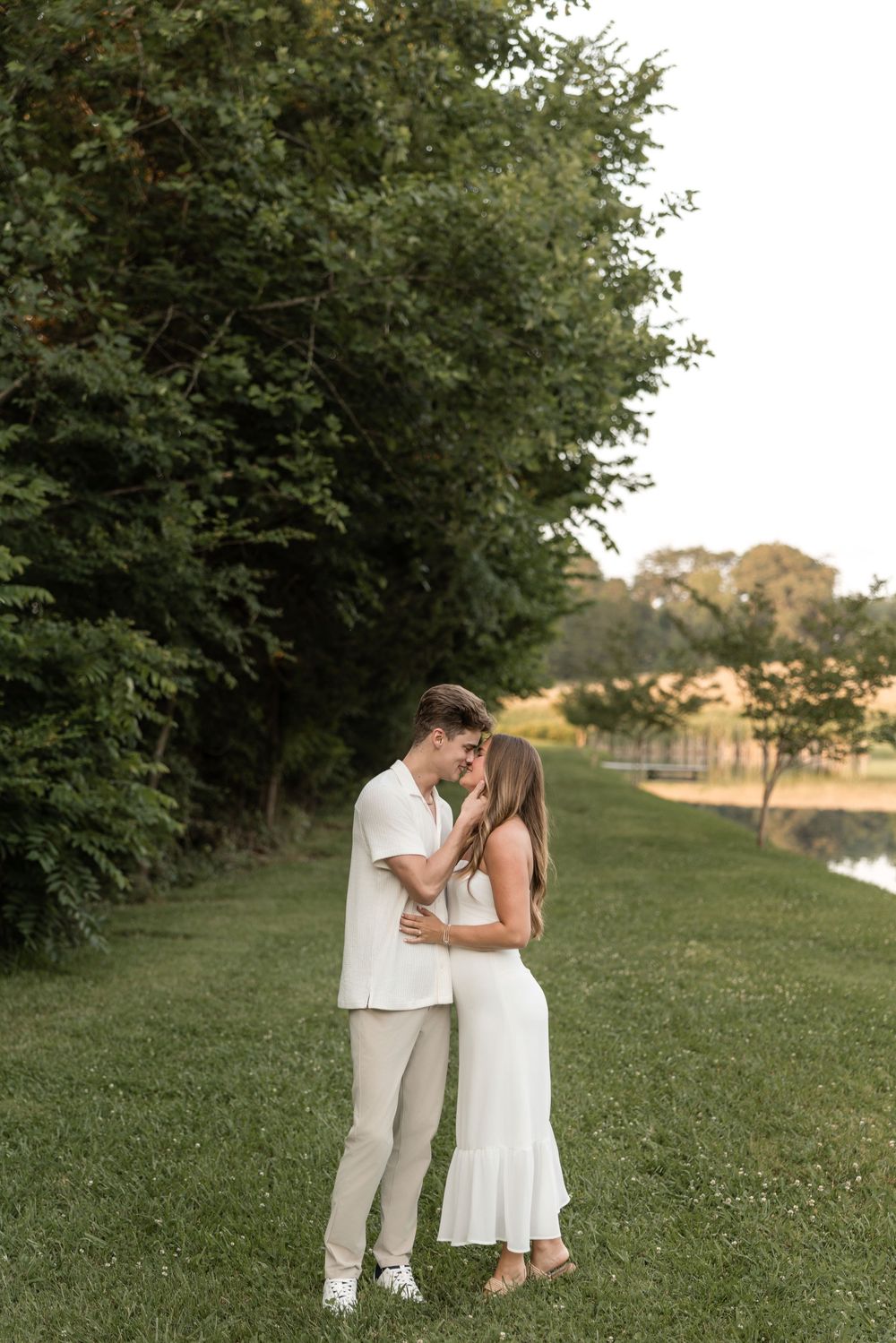A romantic couple in coordinated white outfits embraces on a grassy path next to trees and a pond at sunset.