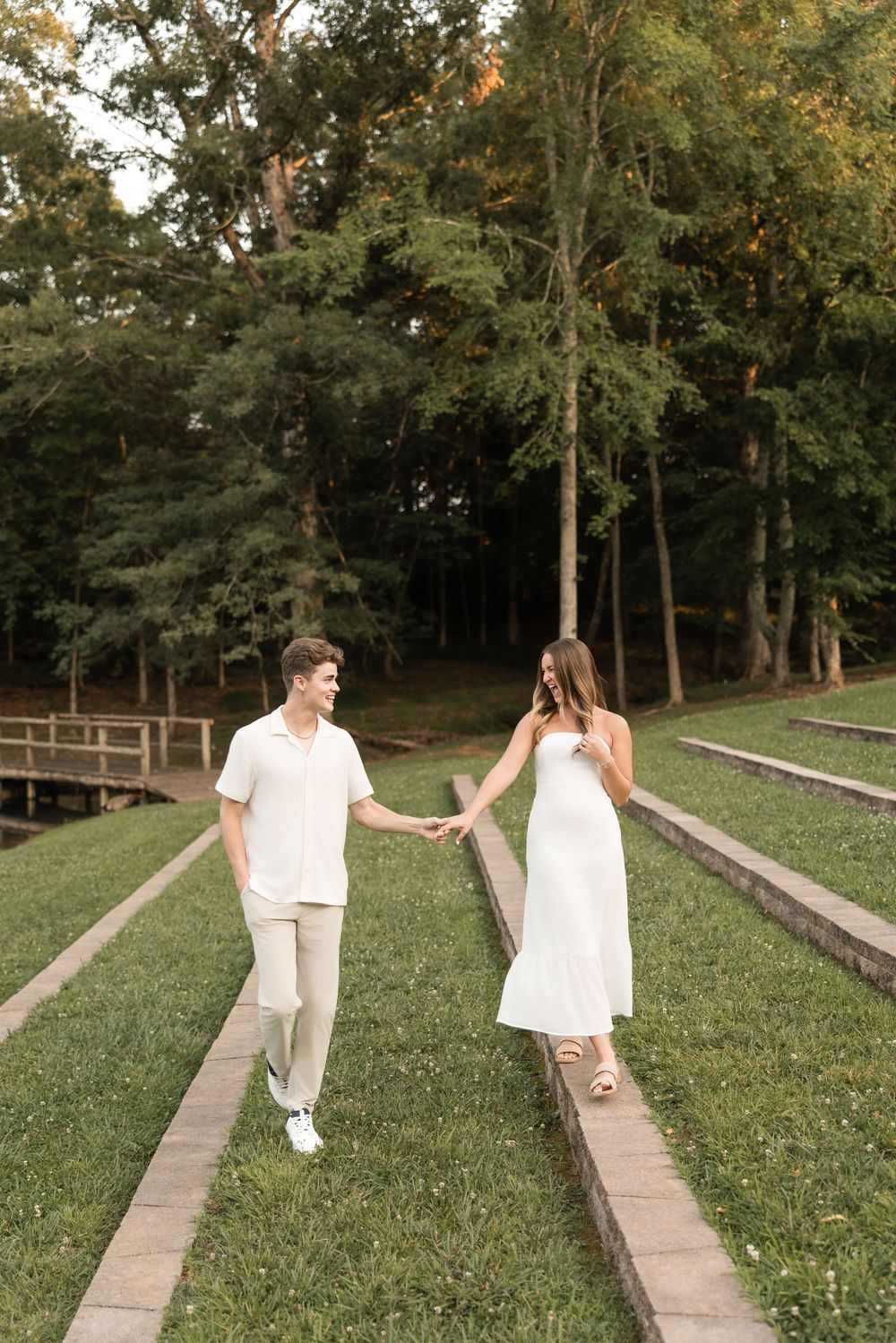 Couple in white outfits walking along parallel garden paths while holding hands in a wooded park setting.
