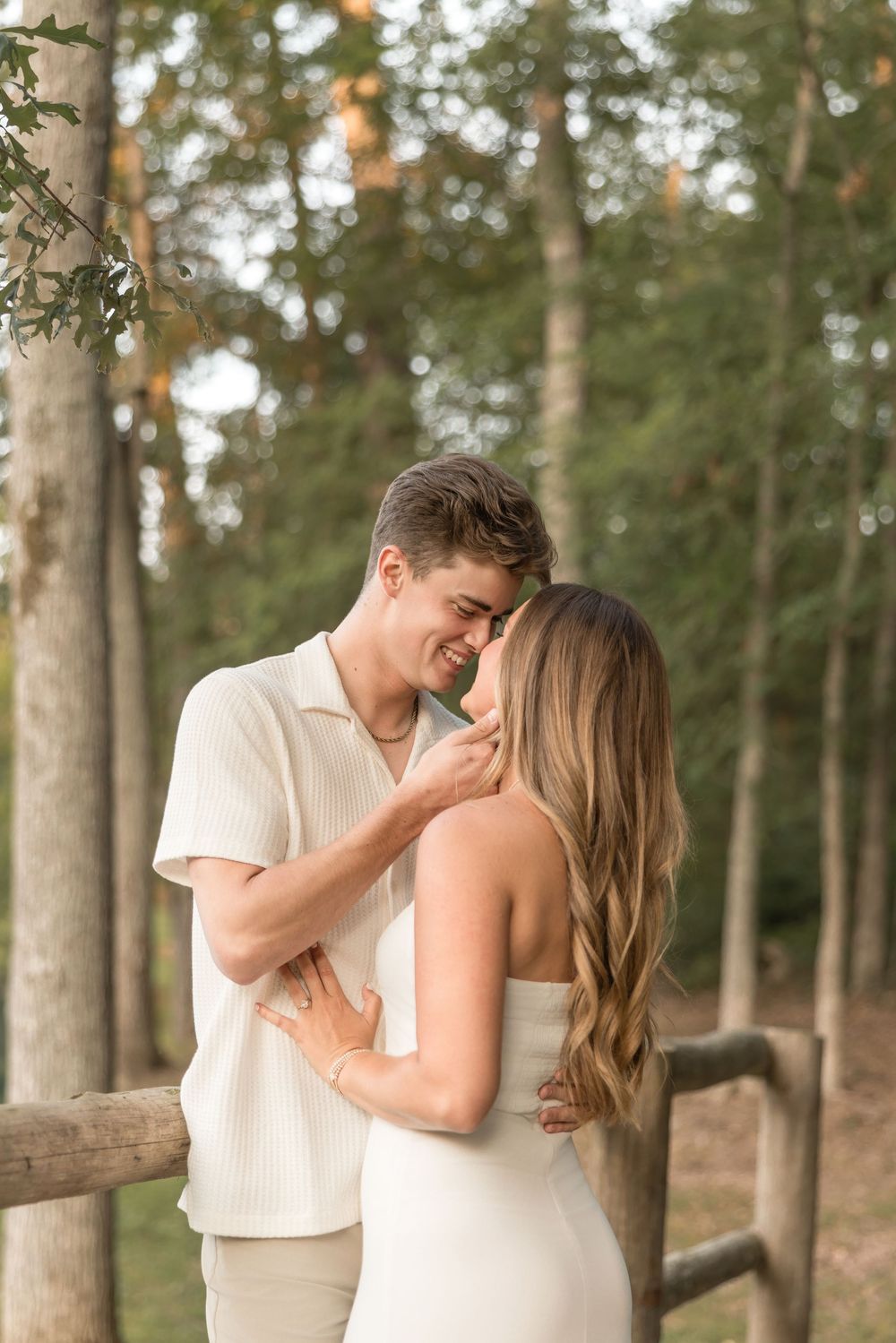 A tender embrace between a couple in white outfits against a wooded forest backdrop.