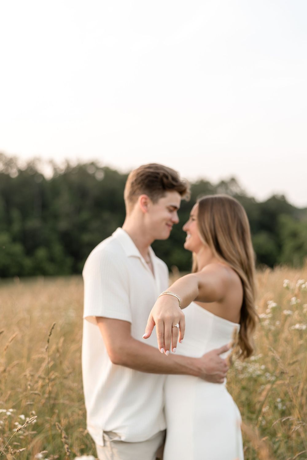 Couple in matching white outfits shows off engagement ring while embracing in a sunlit meadow.