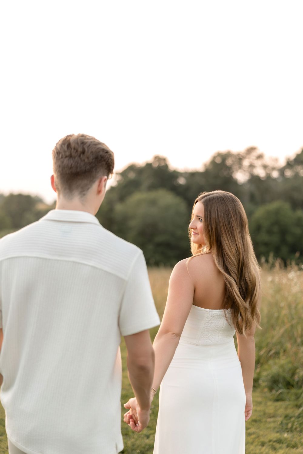 A couple walking through a scenic meadow at sunset wearing coordinating white outfits.