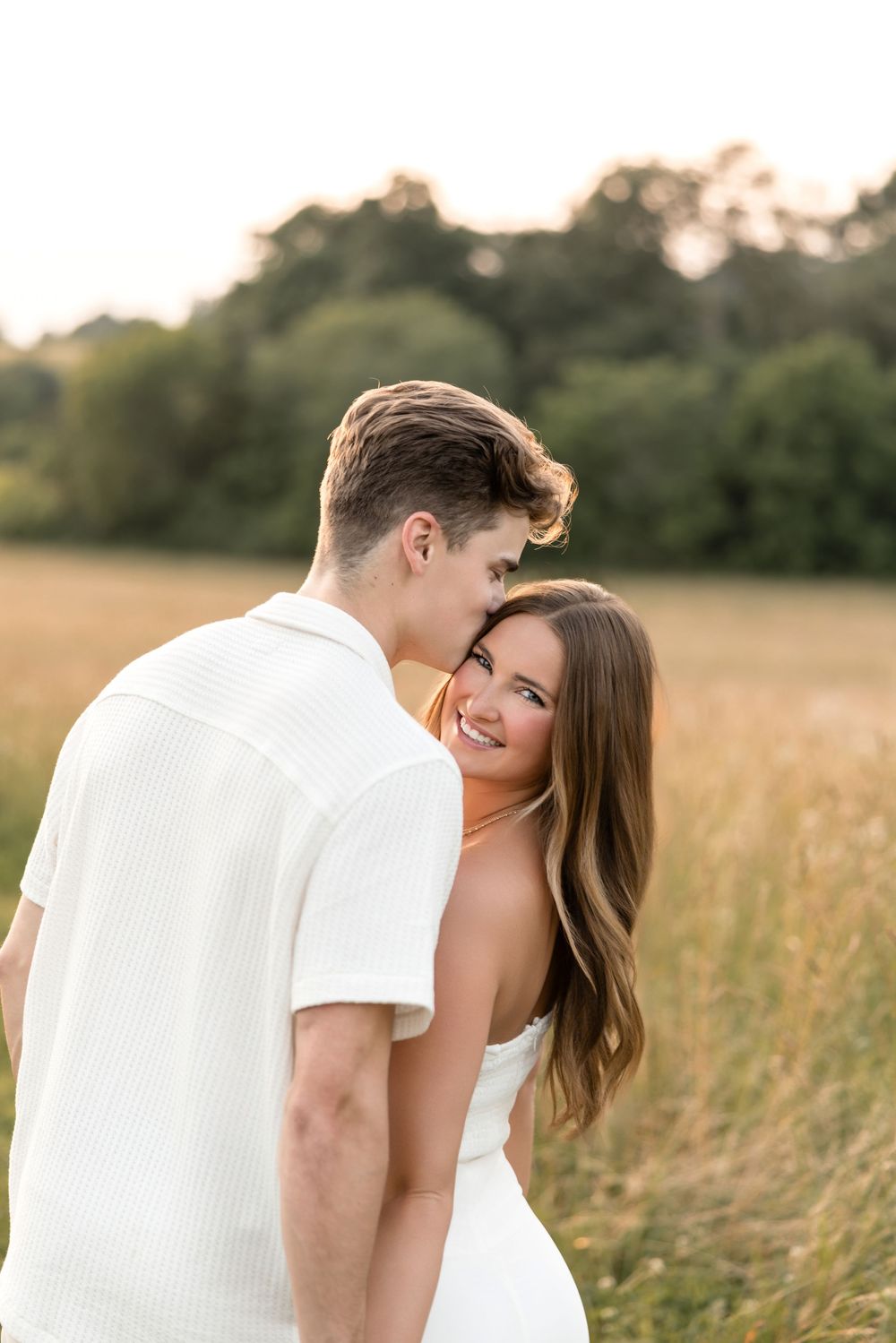 An intimate moment between a couple in white clothing standing in a golden summer field at dusk.