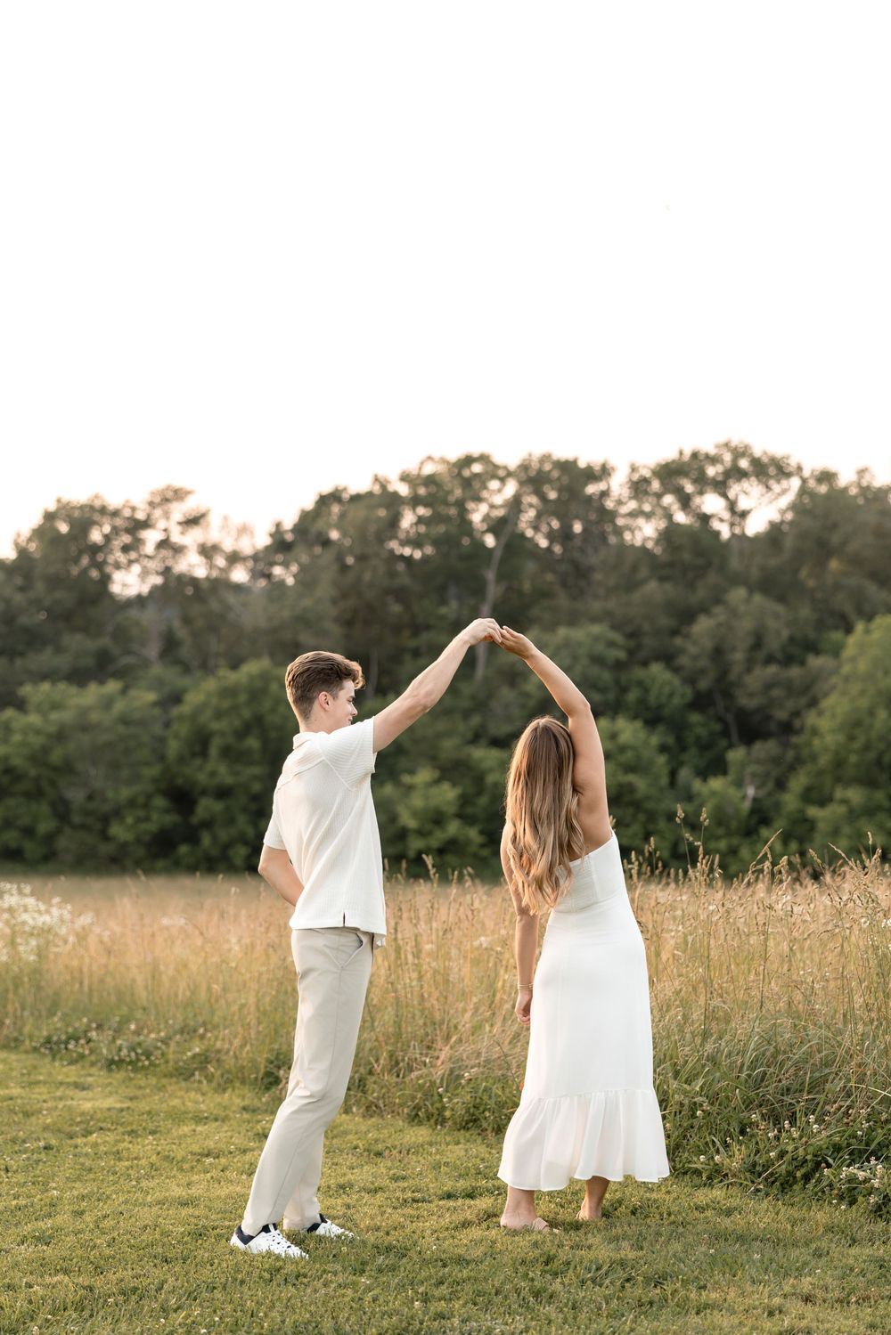 A couple in white outfits dancing playfully together in a golden summer field at sunset.