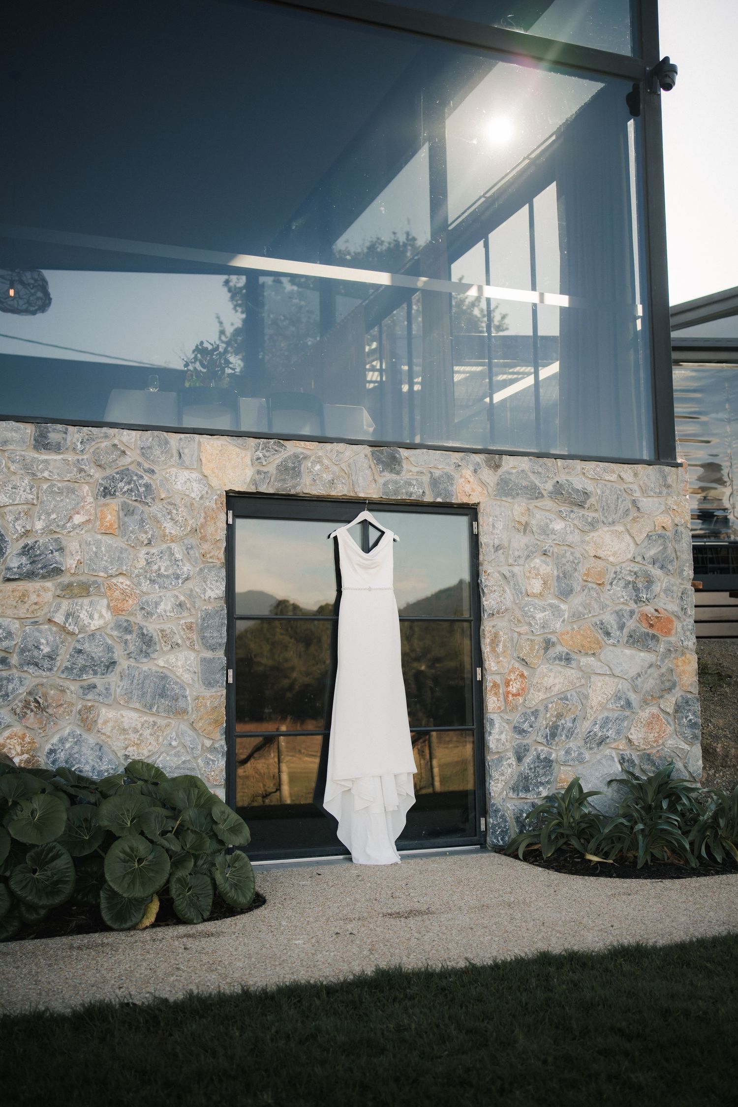 White wedding dress hangs centered in window reflection against stone wall exterior with modern glass architecture above at Boat O Craigo