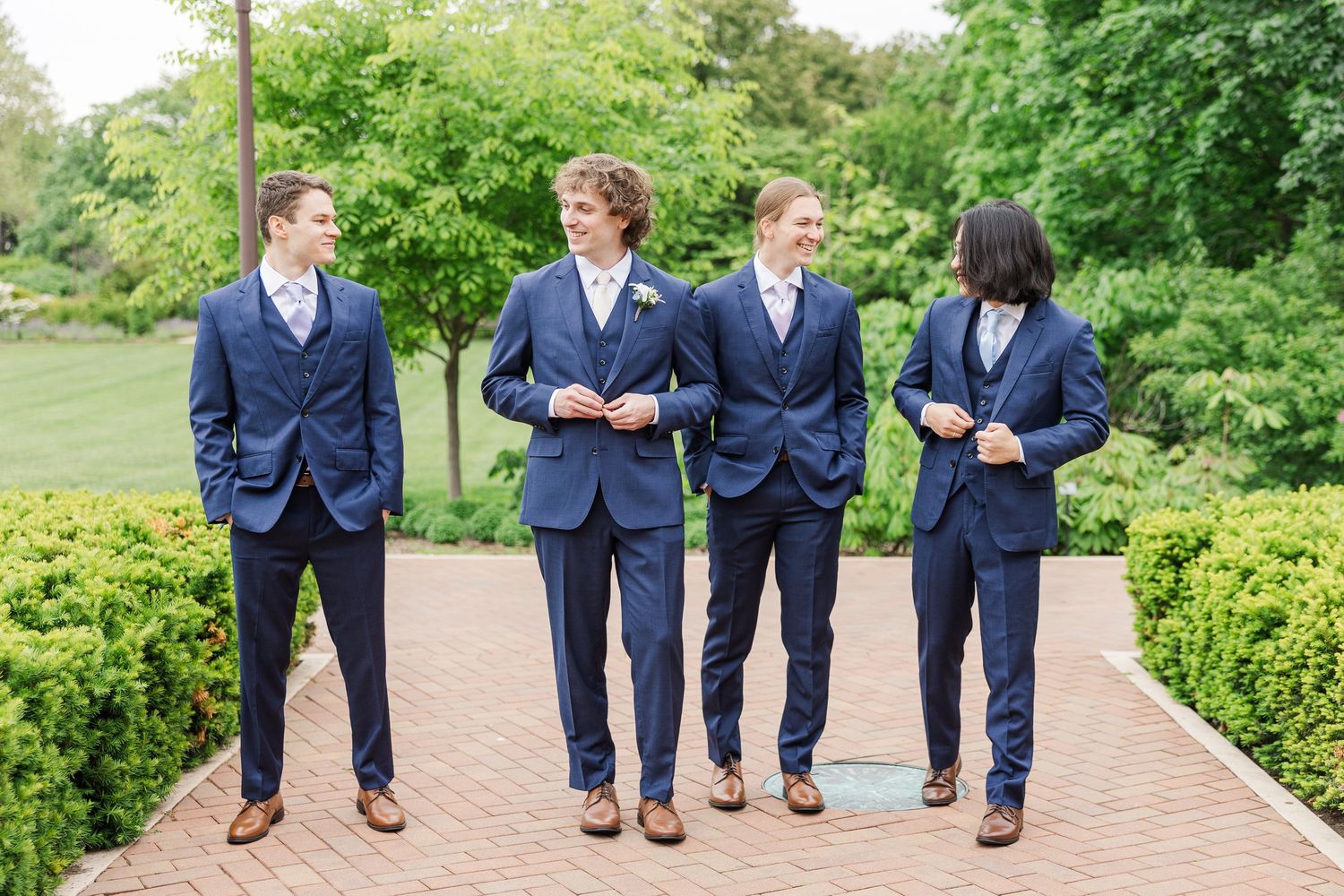 Four friends in matching navy blazers walk and chat together on a brick path surrounded by green garden landscaping.
