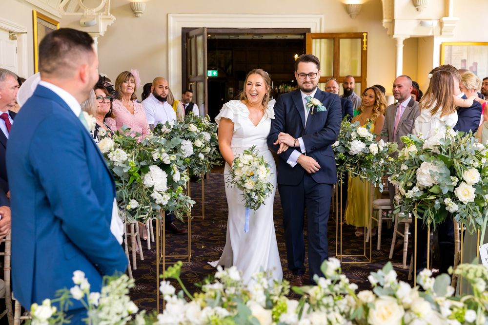 Louisa walking down the aisle with a big smile, surrounded by floral decor at Audley’s Wood Hotel.