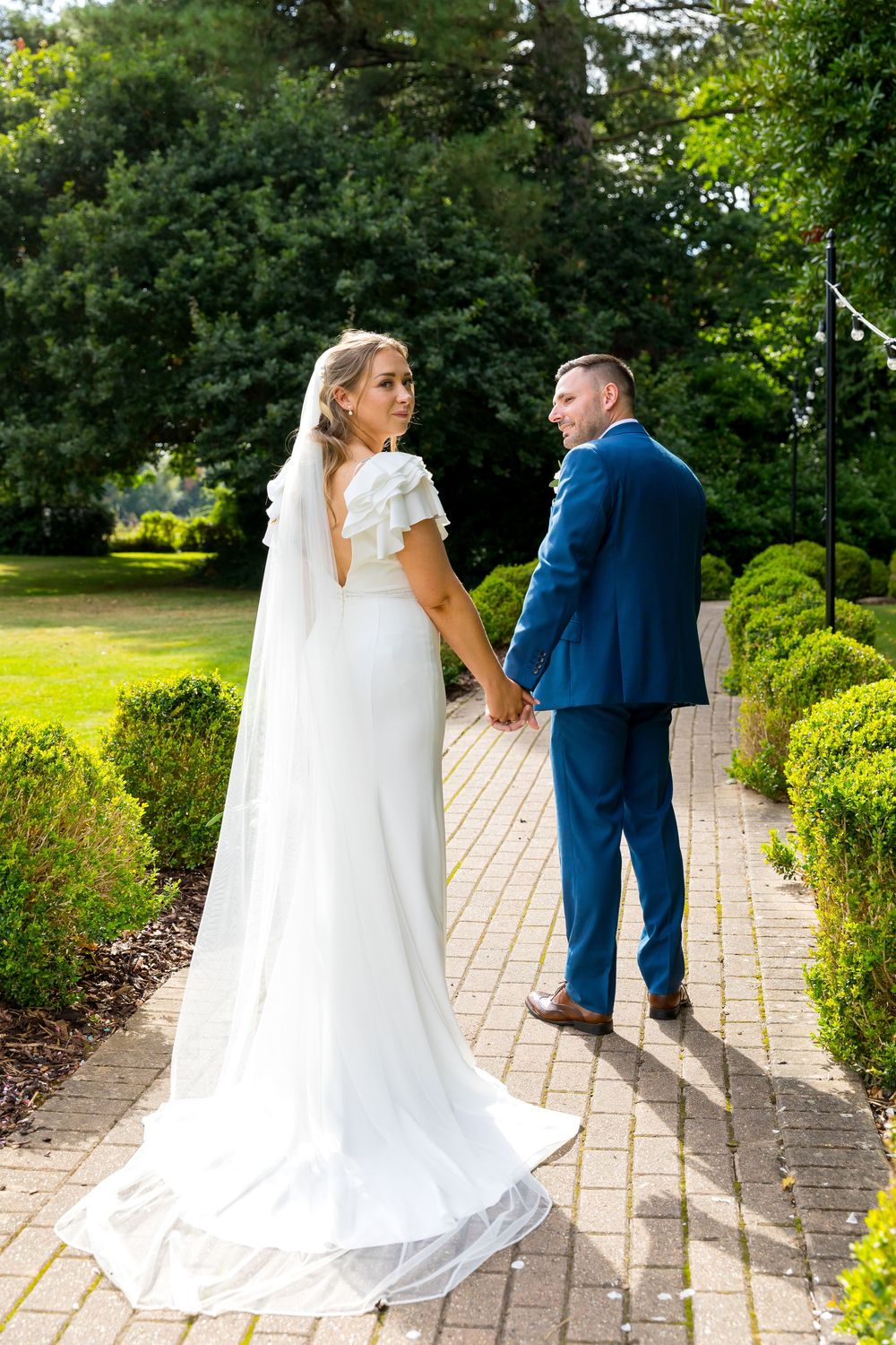 Bride looking back while holding hands with groom on garden walkway