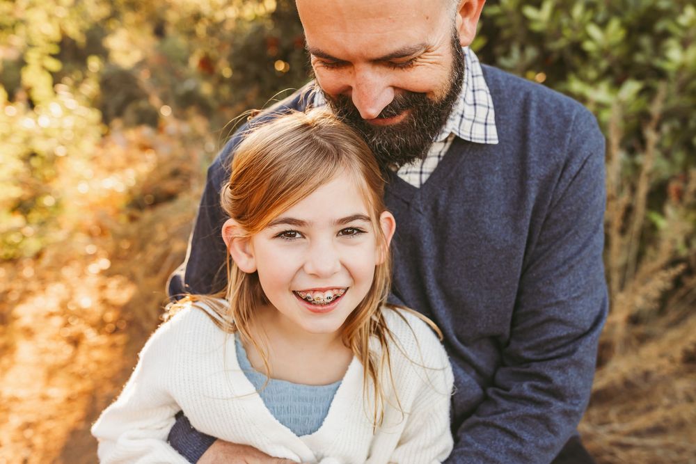 A bearded person in a navy sweater and plaid shirt embraces someone in a playful outdoor portrait at golden hour.