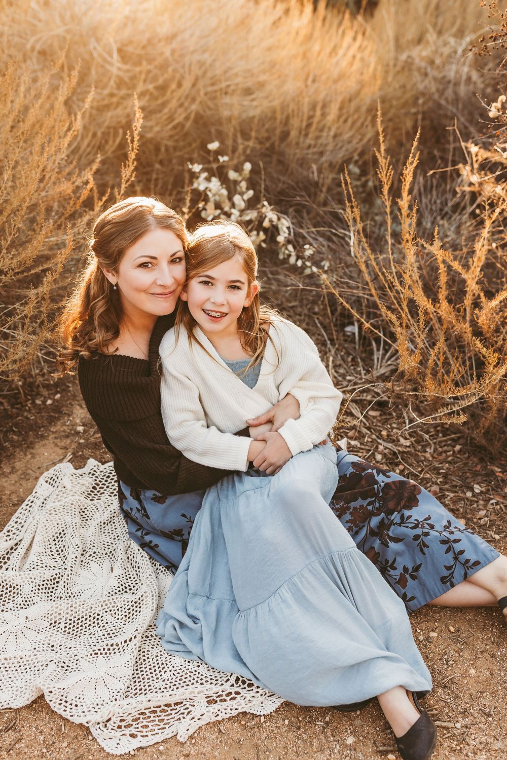 Two people sitting on lace blanket surrounded by golden dried grass during sunset photo session.