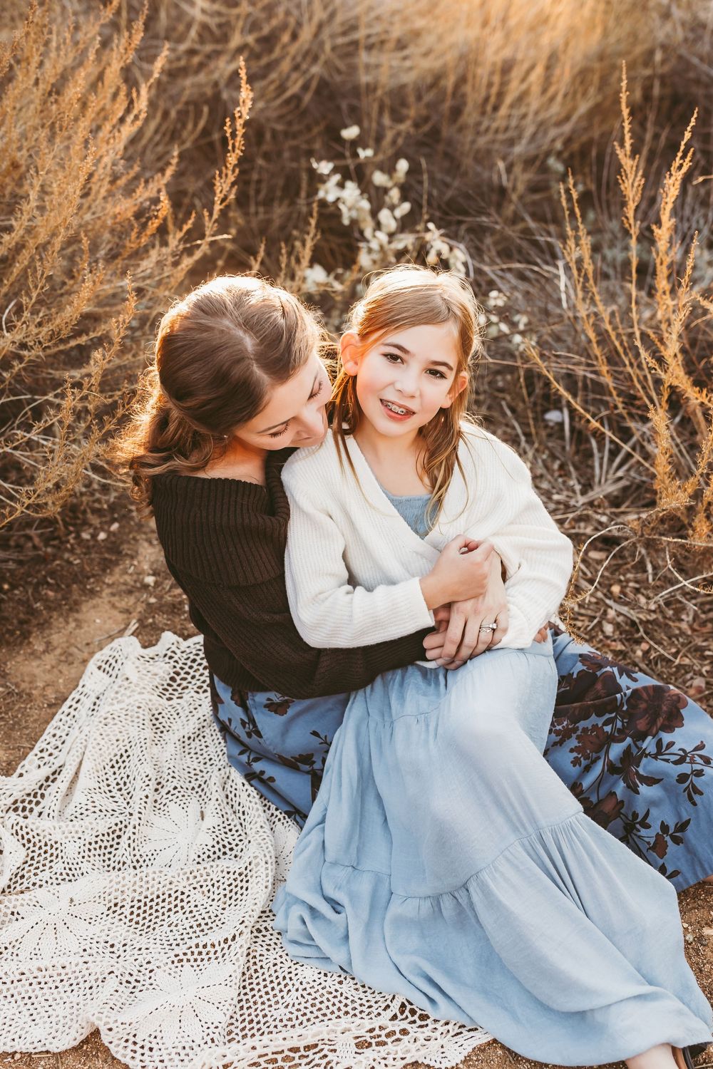 Two individuals sitting together outdoors on a blanket at golden hour with one wearing black and one wearing light blue dress.