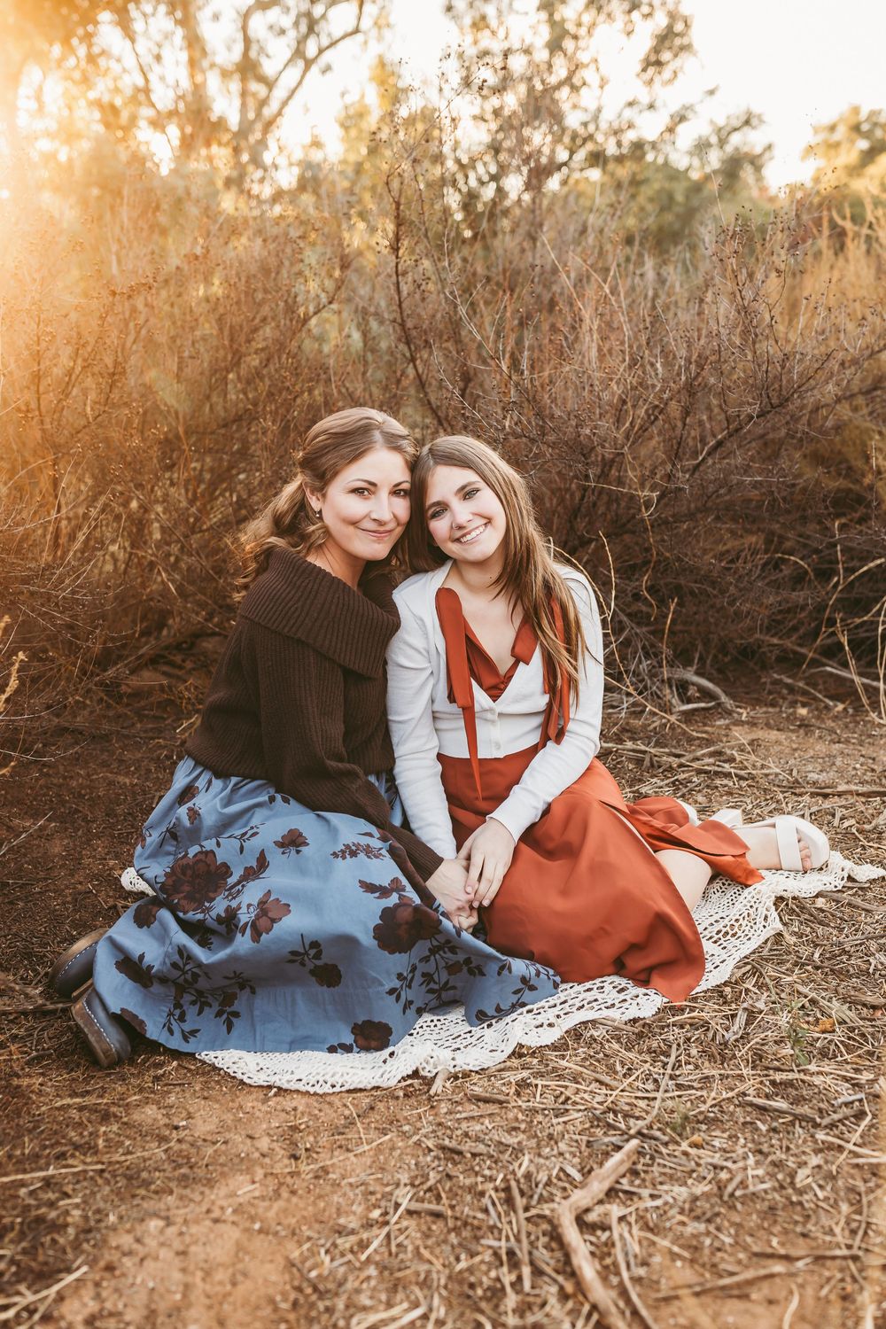 Two people sit together on the ground wearing vintage-style dresses in a sunlit outdoor setting during golden hour.