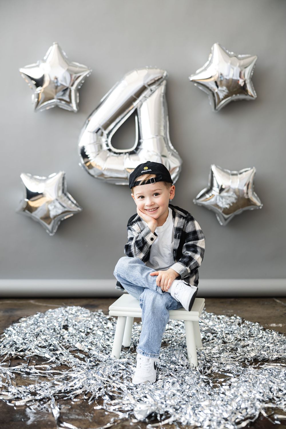Silver mylar number 4 and star balloons decorate a grey wall behind a seated subject with festive decor on the floor.