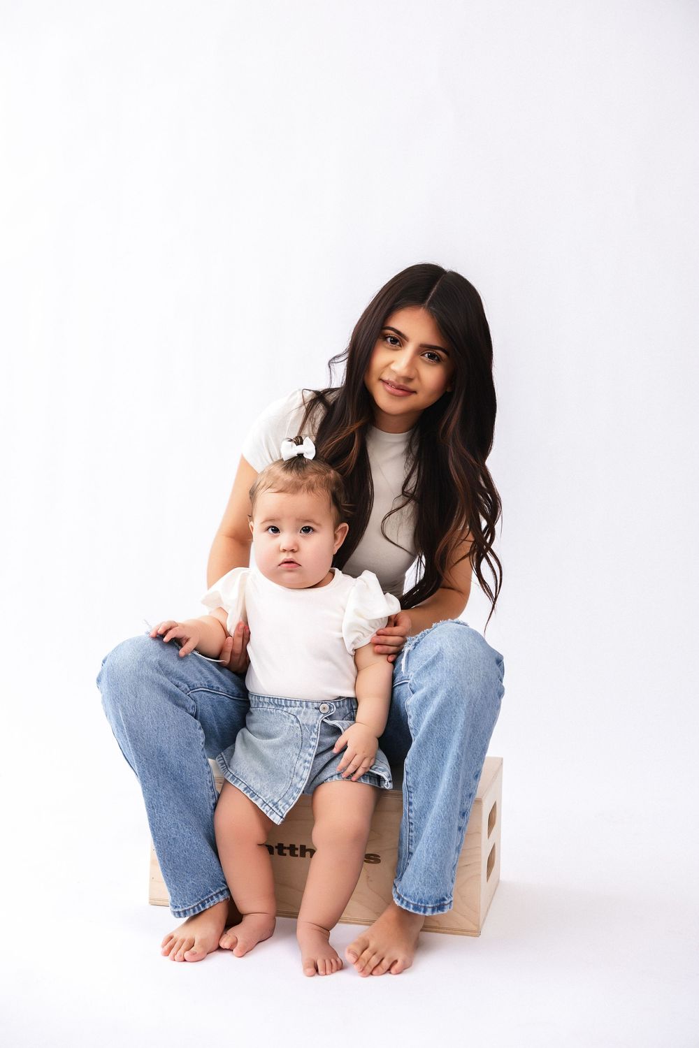 Casual mother and baby sitting barefoot on wooden crate against white background in matching white outfits and denim.