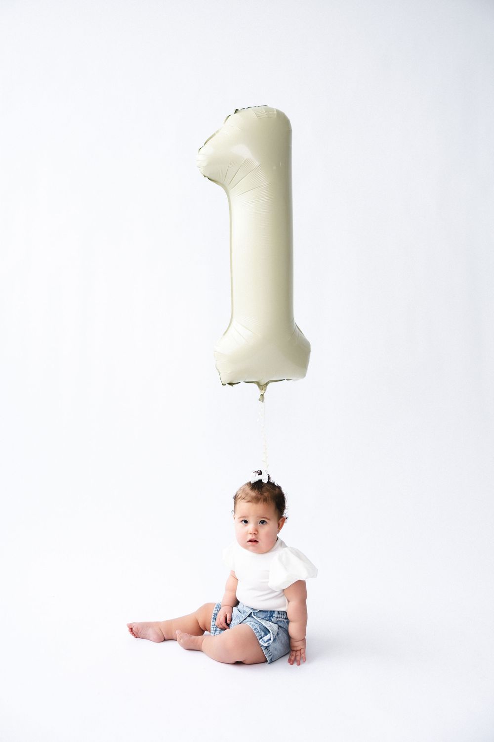 First birthday milestone photo series showing a baby in denim shorts with a giant number one balloon in white against white background.