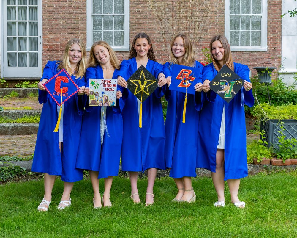 Group in blue graduation gowns hold decorative letters and signs while posing on grass outside brick building.